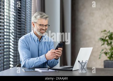 Un uomo d'affari sorridente e maturo che utilizza lo smartphone mentre è seduto alla scrivania con un computer portatile e un notebook. Sembra felice e produttivo. Foto Stock