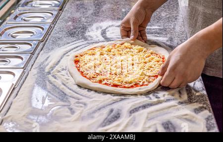 Cuocere in cucina mettendo gli ingredienti sulla pizza. Concetto di pizza. Uomo che aggiunge formaggio alla pizza a tavola, primo piano. Concetto di cucina: Cuocere a mano Foto Stock