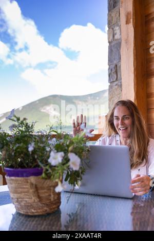 Donna caucasica matura che telelavora con un laptop, in videochiamata situata fuori da una casa all'aperto con montagne sullo sfondo Foto Stock