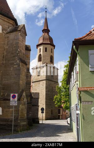 Feuchtwangen, Germania - 6 giugno 2023: Una stretta strada acciottolata si snoda tra edifici in pietra e conduce a un'alta torre della chiesa con un pointe Foto Stock