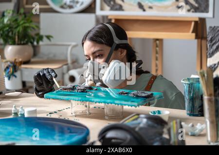 Artista donna seria e concentrata che lavora con resina epossidica, prestando attenzione ai dettagli della pittura. Foto Stock