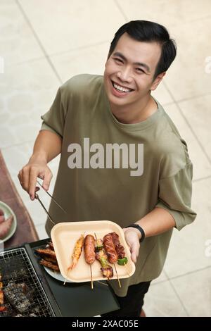 Un giovane sorridente che tiene in mano un piatto con salsicce e gamberetti alla griglia Foto Stock
