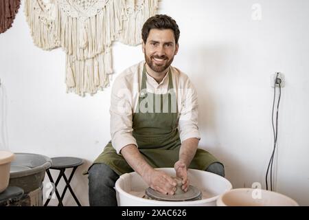 il vasaio maschile in grembiule verde crea piatti in ceramica utilizzando la ruota di un vasaio in un accogliente studio, sorridendo alla macchina fotografica. Foto Stock