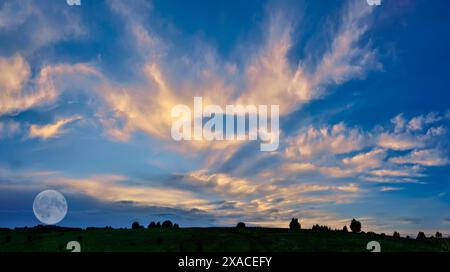 La luna piena sorge sul prato estivo sul meraviglioso cielo del tramonto con nuvole colorate color oro-rosa. Arriva il crepuscolo, la natura si addormenta. Paesaggio estivo al tramonto Foto Stock