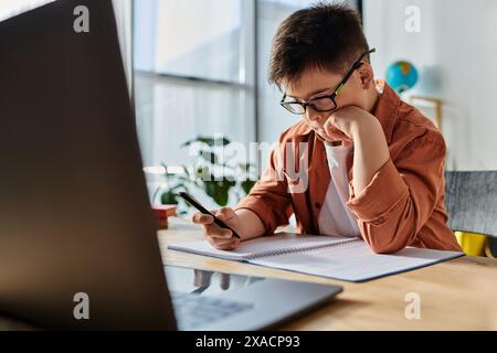 Un ragazzo con la sindrome di Down siede a una scrivania, usando un computer portatile e una penna. Foto Stock