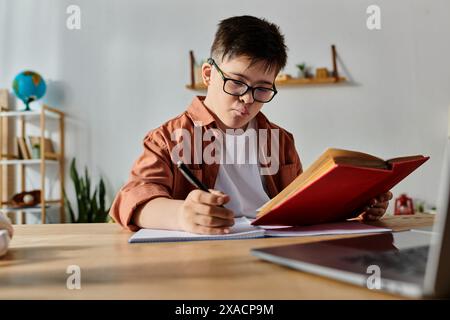 Un ragazzo con la sindrome di Down siede a una scrivania con un portatile e un libro. Foto Stock