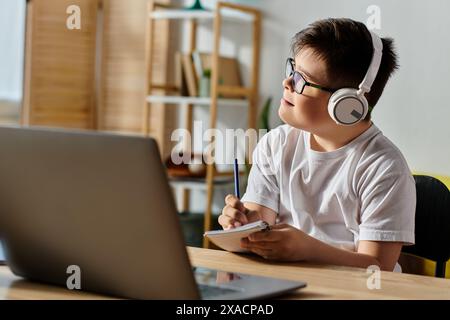 Un ragazzo adorabile con la sindrome di Down che indossa le cuffie siede a una scrivania utilizzando un computer portatile. Foto Stock