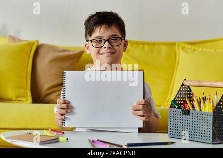 Un adorabile ragazzo con la sindrome di Down mostra con orgoglio un foglio di carta davanti ad un divano giallo. Foto Stock