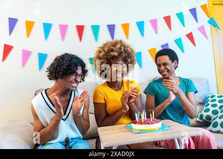 Amici maschi multietnici che si congratulano con una donna africana con una torta di compleanno seduta sul divano sorridendo e applaudendo a casa Foto Stock