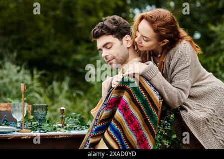 Un uomo e una donna in abiti boho siedono a un tavolo coperto di coperte in un parco verde, godendosi un appuntamento intimo e romantico. Foto Stock