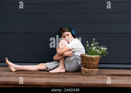 Simpatico ragazzo preadolescente con le cuffie che ascolta la musica su una terrazza di legno vicino a un fiore in un vaso con spazio per copiare Foto Stock