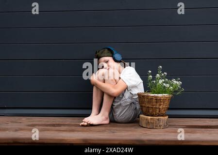Simpatico ragazzo preadolescente in cuffia che ascolta la musica seduto su una terrazza di legno abbracciando le ginocchia Foto Stock