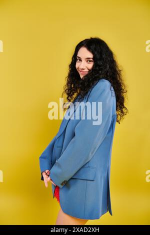 Una giovane donna bruna con capelli ricci, in un abito blu, che posa elegantemente in uno studio con sfondo giallo. Foto Stock