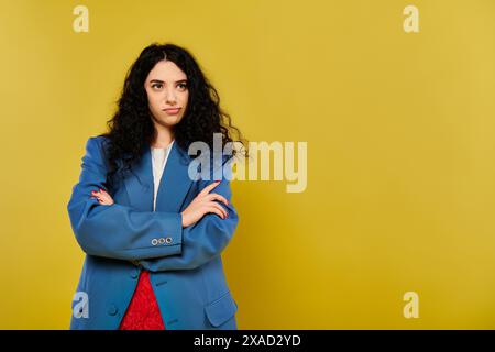 Una giovane donna bruna con i capelli ricci sta con sicurezza con le braccia incrociate di fronte a uno sfondo giallo brillante. Foto Stock