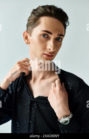 Un giovane con una camicia nera adatta il suo colletto, trasudando sicurezza e stile, in un ambiente da studio su uno sfondo grigio. Foto Stock