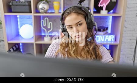 Elegante e giovane donna ispanica, che indossa le cuffie, completamente immersa nel suo videogioco ad alta quota, dal comfort della sua elegante stanza da gioco, Foto Stock