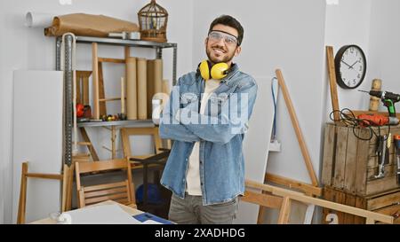 Uomo sorridente con la barba che indossa occhiali di sicurezza e cuffie in piedi con le braccia incrociate in un'officina di falegnameria. Foto Stock