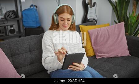 La giovane donna rossa, che indossa le cuffie, ama il tablet digitale sul comodo divano della casa moderna. Foto Stock