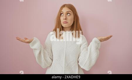 Una giovane donna caucasica con i capelli rossi che indossa un maglione esprime incertezza su uno sfondo rosa Foto Stock