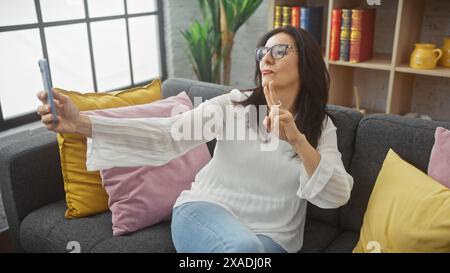 Una donna di mezza età scatta un selfie nel suo salotto moderno, trasmettendo un'atmosfera rilassata e allegra. Foto Stock