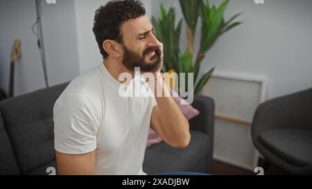 Un uomo barbuto di mezza età che soffre, con la guancia, esprime un mal di denti in un moderno ambiente da salotto. Foto Stock