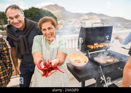 Persone anziane felici che fanno barbecue sul tetto di casa - amici multirazziali che si divertono a mangiare e cucinare insieme durante il giorno del fine settimana - Estate e cibo Foto Stock