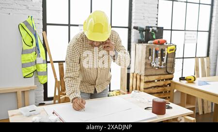 Falegname ispanico in elmetto discutendo i piani al telefono in officina Foto Stock