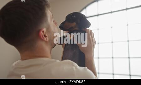Giovane ispanico che tiene in mano un cane da dachshund all'interno in un accogliente soggiorno, che mostra un momento tenero tra l'animale e il suo proprietario. Foto Stock