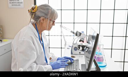 Scienziata matura che lavora con microscopio e computer in laboratorio. Foto Stock