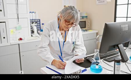 Donna matura con capelli grigi che indossa gli occhiali conduce ricerche in laboratorio, scrivendo appunti. Foto Stock