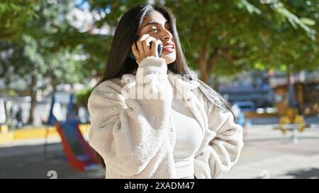 Una giovane donna indiana con i capelli lunghi parla al telefono in un parco soleggiato, irradiando bellezza urbana in mezzo alla natura. Foto Stock