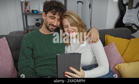 Una coppia amorevole si siede da vicino su un divano in un moderno soggiorno, condividendo un tablet e divertendosi insieme. Foto Stock