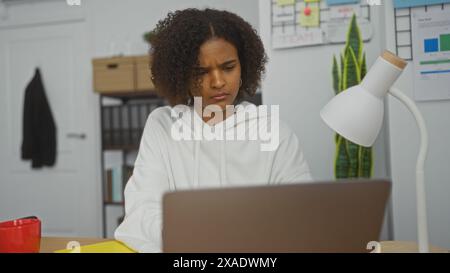 Giovane donna afroamericana con capelli ricci che lavora in casa in un ufficio, concentrandosi con attenzione sul suo laptop, con una lampada, documenti e un rosso Foto Stock