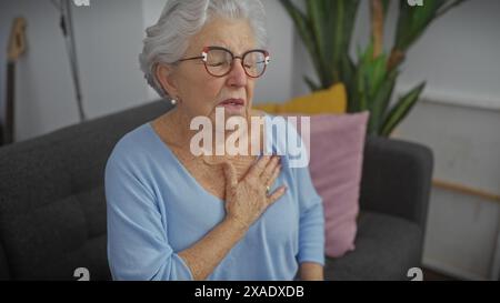 Donna anziana con occhiali che soffre di disagio in salotto, toccando il petto. Foto Stock