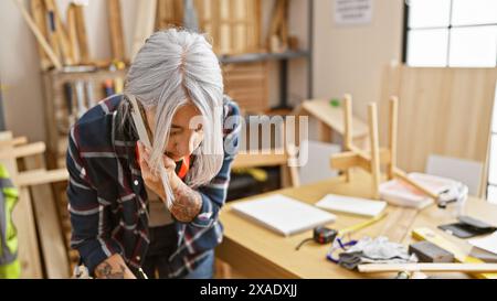 Un'affascinante carpentiere di mezza età dai capelli grigi parla con passione sullo smartphone in mezzo alla segatura nel suo vivace studio di falegnameria al chiuso Foto Stock