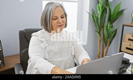 Una donna matura lavora con attenzione al suo notebook in un ambiente moderno, incarnando professionalità e concentrazione. Foto Stock