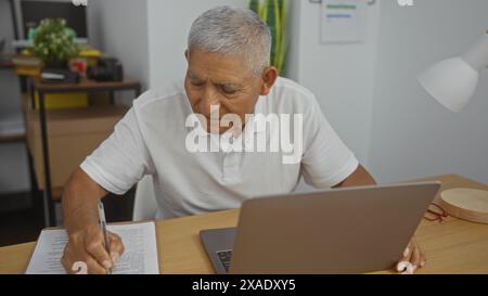 Uomo ispanico dai capelli grigi che prende appunti in un ufficio, seduto a una scrivania con un laptop, in un moderno ambiente di lavoro al chiuso. Foto Stock