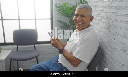 Un bell'uomo ispanico dai capelli grigi siede in una luminosa stanza interna sorridente e regge un telefono, con un muro di mattoni bianchi e una finestra sullo sfondo Foto Stock