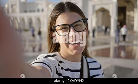 Una donna sorridente con gli occhiali scatta un selfie al qasr al watan di abu dhabi, emirati arabi uniti, mostrando turismo e architettura. Foto Stock