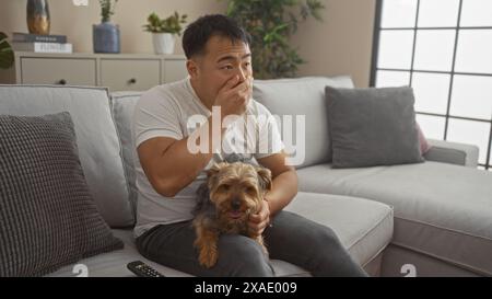 Un giovane cinese è seduto al chiuso su un divano in un salotto mentre accarezza il suo cane, apparendo sorpreso mentre guarda qualcosa con attenzione. Foto Stock