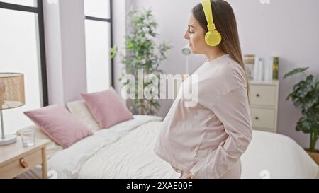 Una donna incinta con le cuffie sta in piedi in modo contemplativo nella sua moderna camera da letto, incarnando tranquillità e aspettativa. Foto Stock