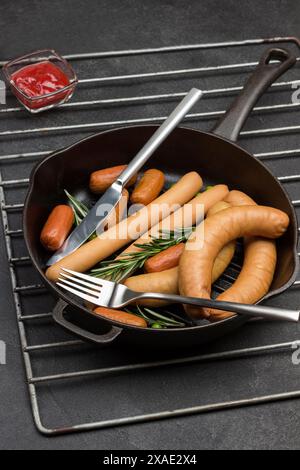 Salsicce e ciuffi di rosmarino con coltello e forchetta in padella su griglia metallica. Pasta di pomodoro, senape e rafano in tavola. Sfondo nero. T Foto Stock