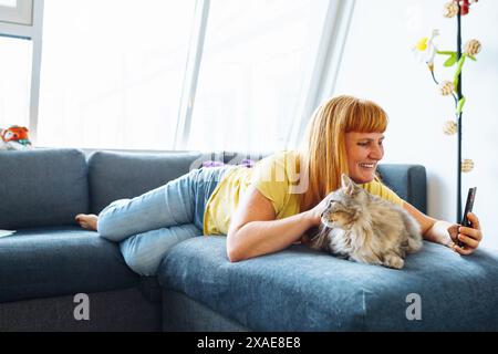 ritratti una donna bionda di mezza età sdraiata sul divano abbracciando un gatto che scatta selfie Foto Stock