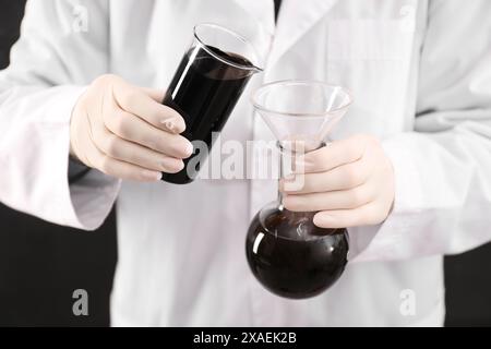 Donna che versa olio grezzo nero dal becher al matraccio su sfondo scuro, primo piano Foto Stock