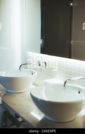 Un bagno moderno con due lavandini in ceramica bianca montati su un ripiano in marmo, con rubinetti cromati e uno specchio ben illuminato su una t Foto Stock