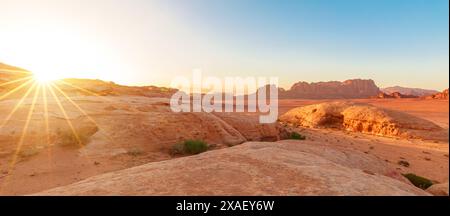 panorama del tramonto nel deserto con il sole che tramonta dietro le aspre montagne di Wadi Rum, bagnando il vasto deserto giordano in calde tonalità dorate sotto a. Foto Stock