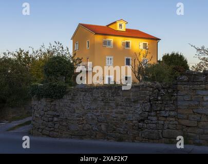 Foto di una casa gialla con balcone bianco, finestre e tetto rosso circondati da giardino e pareti durante il tramonto estivo Foto Stock