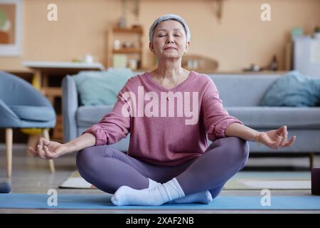 Ritratto a figura intera di una donna anziana dai capelli grigi che pratica yoga a casa e medita con gli occhi chiusi in uno spazio copia in posizione loto Foto Stock