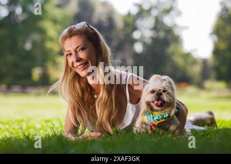 Una donna bionda allegra gode di un momento di pace con il suo piccolo cane in un parco soleggiato, entrambi con espressioni felici. Foto Stock