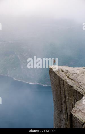 Un escursionista solitario si trova ai margini del Preikestolen, una spettacolare scogliera in Norvegia. Il paesaggio nebbioso e la goccia sottostante evocano un senso di soggezione e solitudine. Foto Stock